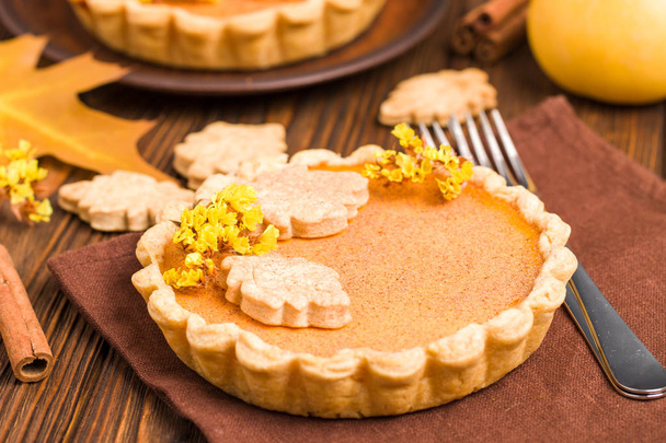 Pumpkin pie with cinnamon and cookies on brown napkins on wooden background with autumn yellow leaves - top view close up photography of seasonal american traditional sweet baked food. - Photo, Image