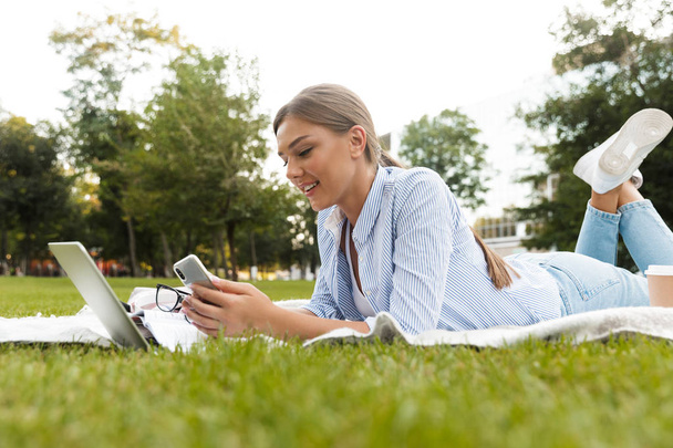 Sorrindo jovem passando tempo no parque, estudando, deitado em um cobertor com computador portátil, usando telefone celular
 - Foto, Imagem