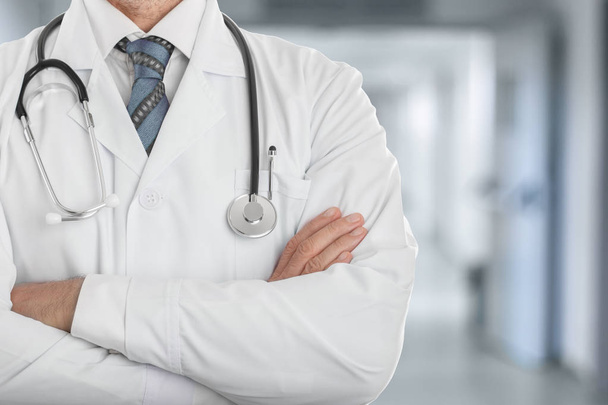 Médico masculino de casaco branco com estetoscópio em pé com as mãos cruzadas na clínica
 - Foto, Imagem