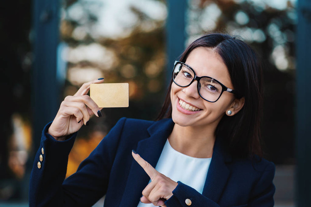 Successful pretty woman look to camera and showing unlimited gold credit card on office building background. Professional female manager wearing glasses and suit - Foto, afbeelding