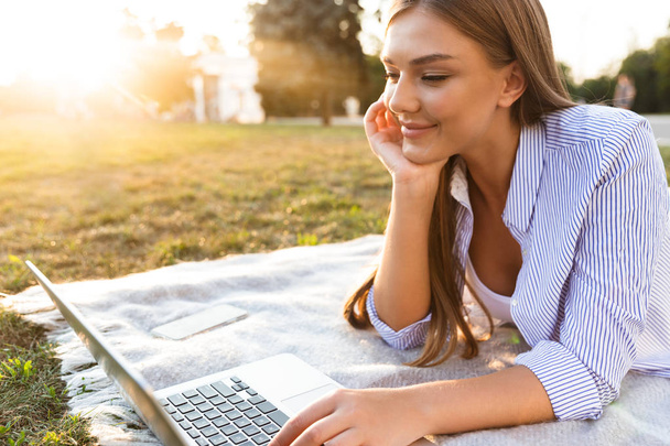 Pretty young girl spending time at the park, studying, laying on a blanket with laptop computer - 写真・画像