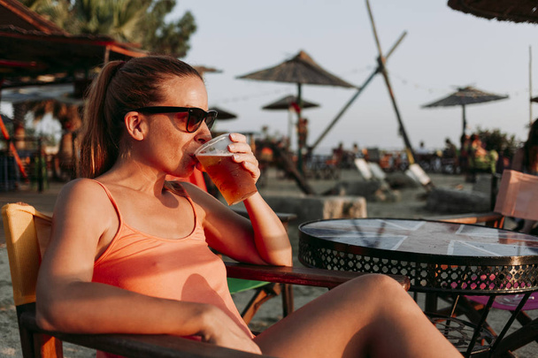 Young woman drinking beer in a beach bar enjoying sunset - Photo, Image
