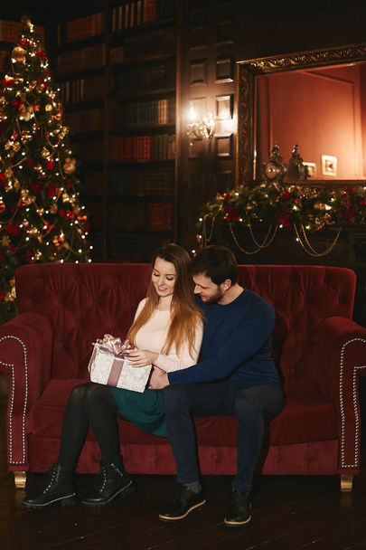 Fashionable and beautiful couple, sexy brunette model girl with bright makeup in a trendy skirt with a gift in hands and stylish handsome bearded men, sits on a sofa and posing at Christmas interior - Photo, image