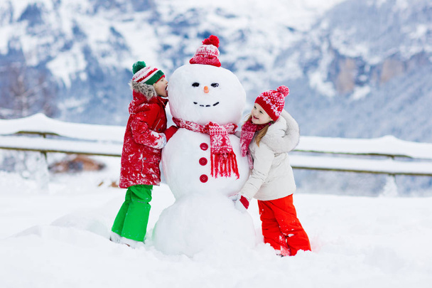 Un bonhomme de neige. Les enfants construisent un bonhomme de neige. Garçon et fille jouant dehors le jour d'hiver enneigé. Amusement en famille en plein air pendant les vacances de Noël dans les montagnes. Les enfants jouent dans le paysage montagneux suisse. - Photo, image