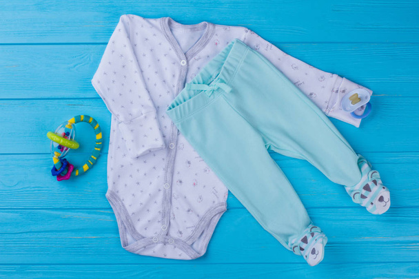 Bebek pijama ve rattle oyuncak. Beyaz toddler onesie ve komik ayaklı pantolon. Mavi ahşap arka plan. - Fotoğraf, Görsel