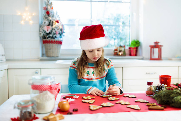 Kids bake Christmas cookies. Child in Santa hat cooking, decorating gingerbread man for Xmas celebration. Family preparing sweets in white kitchen with Christmas tree on snowy winter day. - Photo, Image