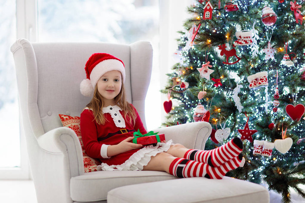 Lapsi koristelu joulukuusi ja avaaminen läsnä ja lahja kotona. Joululahjoja ja leluja. Pikkutyttö joulupukin puvussa ja hattu takassa. Perhe lasten kanssa juhlia talvilomaa
. - Valokuva, kuva