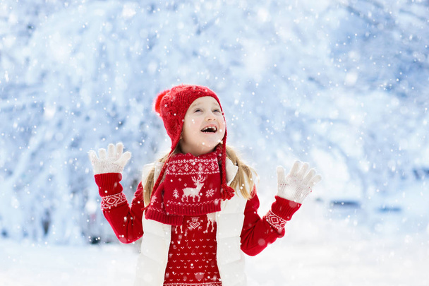 Child in red hat playing in snow on Christmas vacation. Winter outdoor fun. Kids play in snowy park on Xmas eve. Little girl in knitted sweater, scarf and mittens with Christmas decoration. - Foto, Imagem