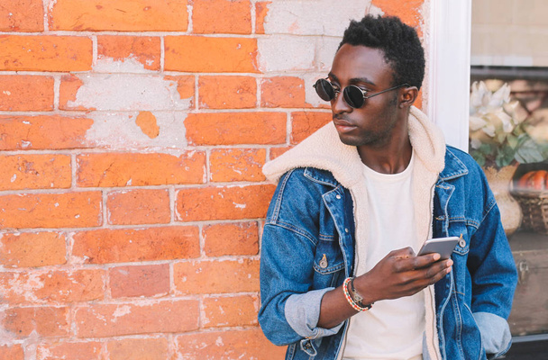 Fashion african man holds smartphone on city street, brick wall background - Foto, immagini