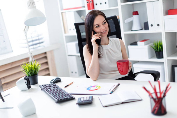 ein junges Mädchen sitzt im Büro am Computertisch, telefoniert und hält eine rote Tasse in der Hand. - Foto, Bild
