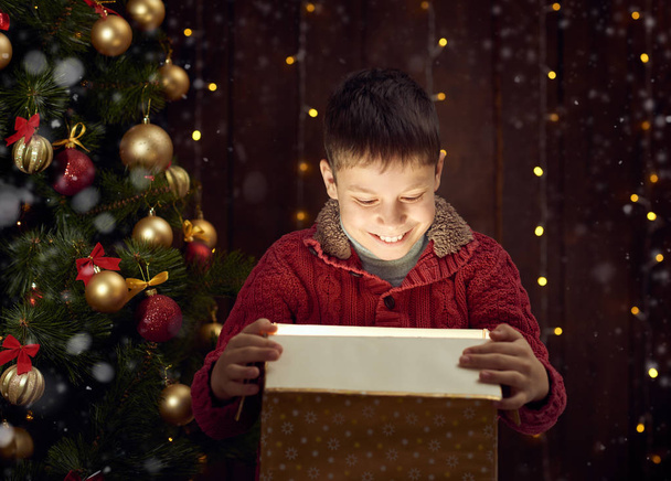 lapsi poika istuu lahjapakkauksessa lähellä joulukuusi koristeltu kuusen, tumma puinen tausta
 - Valokuva, kuva