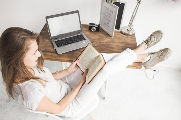 Young business woman working at home and reading a book with her legs on the table. Creative Scandinavian style workspace. Wiew from above - Фото, изображение