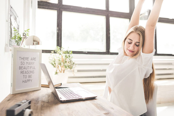 Jovem mulher de negócios trabalhando em casa atrás de um laptop e esticando as mãos. Espaço de trabalho estilo escandinavo criativo
 - Foto, Imagem
