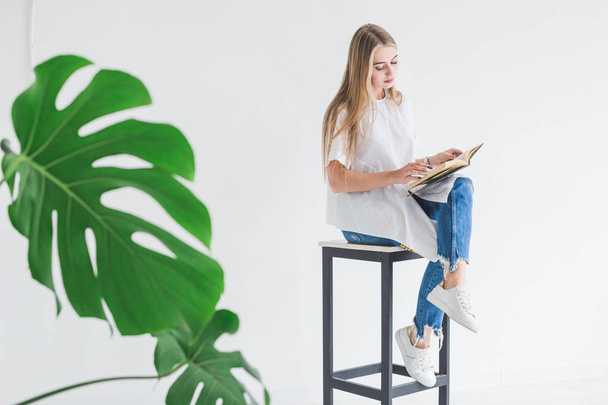 Portrait of a young stylish blonde girl in a white T-shirt and blue jeans reading a book on a white background - Fotó, kép