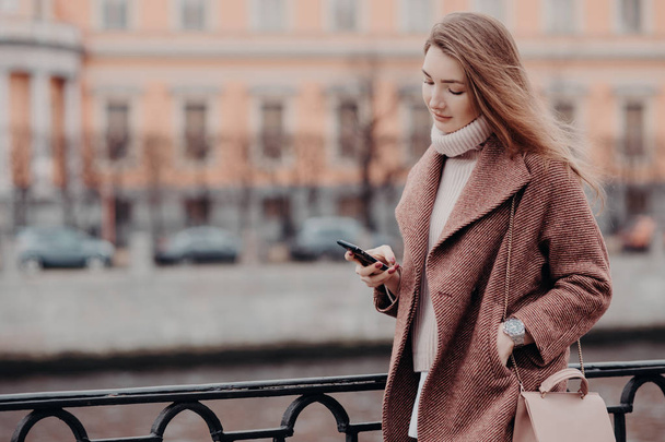 Mooie vrouw in warme jas, houdt moderne mobiele telefoon, berichten in sociale netwerken, verbonden met gratis wifi, wandelen in de stad, street style, onscherpe achtergrond met kopie ruimte voor uw advertentie - Foto, afbeelding