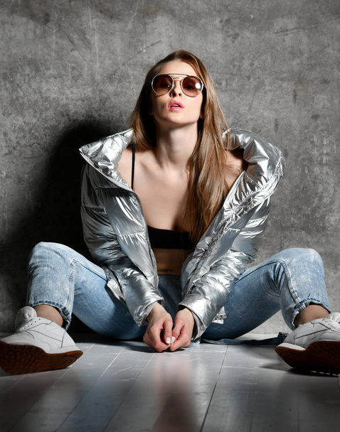 sport woman after workout exercise posing in silver shiny puffer jacket blue jeans outwear sunglasses on loft wall   - 写真・画像
