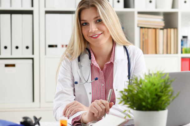 Όμορφη χαμογελαστή γυναίκα γιατρό να καθίσει στο χώρο εργασίας - Φωτογραφία, εικόνα