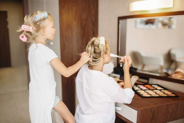 Child makes a funny makeup to her mother against a mirror in bedroom at home. Parent feeling, togetherness, happy times - Foto, Imagen