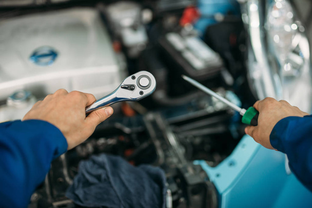 Male technician hands with screwdriver and wrench, car with opened hood on background. Auto-service, vehicle maintenance - Photo, Image