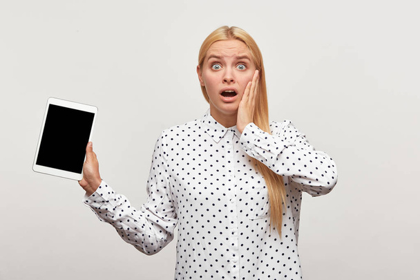 Emotional blonde girl with wide open eyes and mouth, looks shocked with tablet in hand, one hand holding her cheek, black screen looks camera for your message or advertisement, on white background
 - Фото, изображение