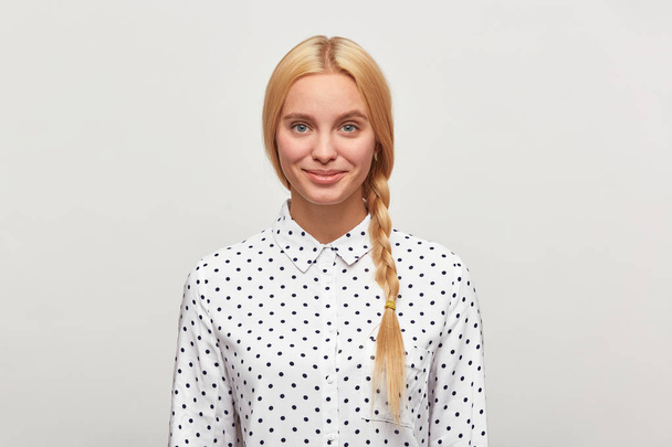 Beautiful diligent blonde woman with blue eyes, braid, feels happy, tenderly smiles, wears white shirt with black polka dots. Modest, excellent student, came to interview. Over white background - Foto, Bild