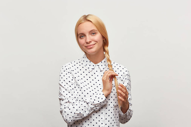 Beautiful diligent blonde woman with blue eyes, corrects keeps braid, feels happy, shows tenderness, wears white shirt with black polka dots. Over white background - Photo, Image