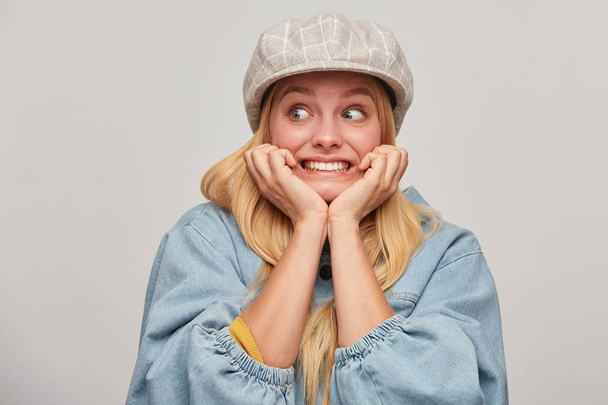 Scared blonde girl looks aside frightened afraid chattering teeth with fear,bites nails from fright, hands holding cheeks, wears oversize denim jacket and beige checked cap,on grey background - Photo, Image