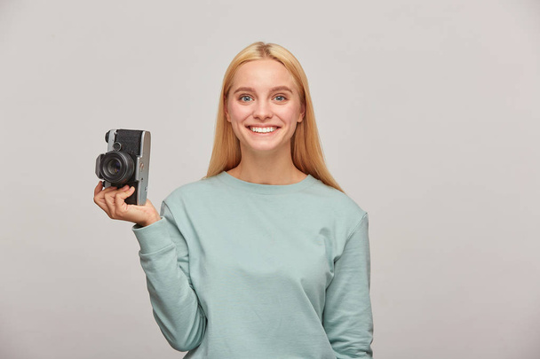 schöne Fotografin sieht glücklich lächelnd aus, hält eine Retro-Vintage-Fotokamera in der einen Hand, genießt den Shootingvorgang, trägt blaues lässiges Sweatshirt, vor grauem Hintergrund - Foto, Bild