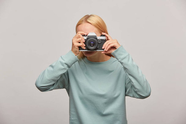 Menina cobrindo seu rosto com a câmera fazendo uma sessão de fotos. Fotógrafo tirando foto, trabalhando em estúdio, fotografia na porta, camisola azul vestida, sobre fundo cinza
 - Foto, Imagem