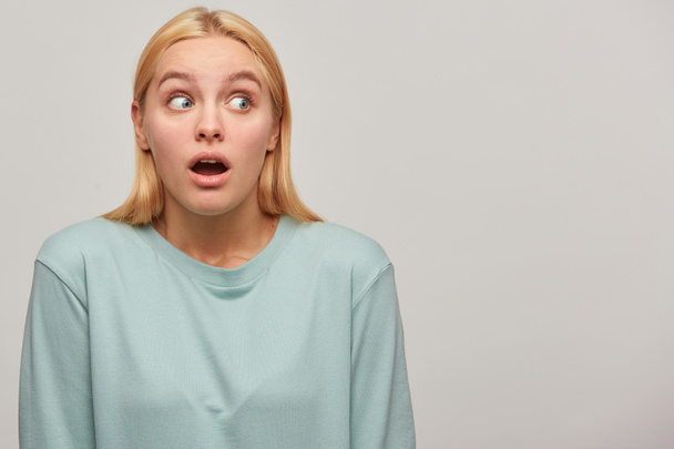 Worried scared blonde girl looks aside around with mouth opened, frightened afraid, presses down her head in shoulders, wearing blue casual sweatshirt, copyspace on the right, grey background - Photo, Image