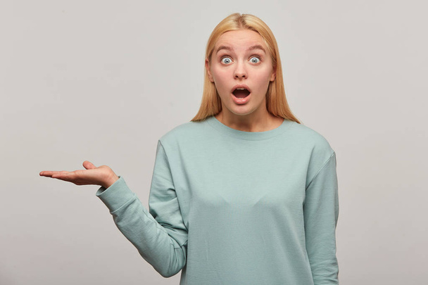 Shocked blonde young girl, looking wonderstruck amazed open-eyed wide open mouth, holding the copyspace on one palm, wearing blue casual sweatshirt,isolated over grey background - Photo, Image