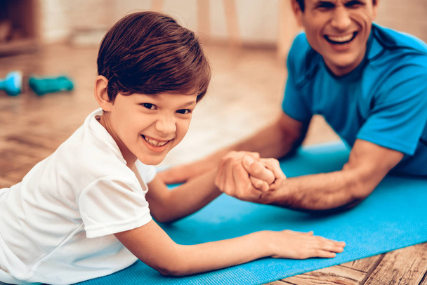 Boy Standing on Scales. Father and Son do Spotting. Sport at Home. Warm Up in Quarter. Lying on Gymnastic Mat. Dumbbells in Hands. Boxing Gloves. Doing Sports. Man and Boy Train at Home. Swing Press.  - Photo, image