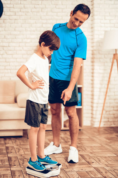 Boy Standing on Scales. Father and Son do Spotting. Sport at Home. Warm Up in Quarter. Lying on Gym Mat. Dumbbells in Hands. Boxing Gloves. Doing Sports. Man and Boy Train at Home. Stand on Scales. - Photo, Image