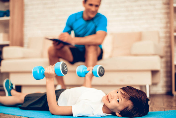 Sports for Kids. Boy with Dumbbells in Hands. Boy with Dumbbells in Hands. Father and Boy Do Spotting. Sport at Home. Health Concept. Healthy Lifestyle. White Sofa. Lying on Floor. Male Trainer. - Foto, afbeelding