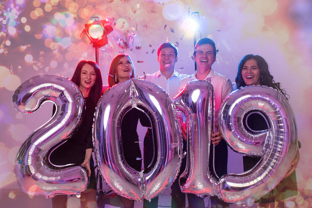 Nouvel an 2019 arrive. Groupe de jeunes gens joyeux tenant des numéros de couleur argent et jetant confettis sur la fête
. - Photo, image