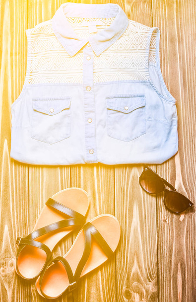 Καλοκαίρι Γυναικεία ρούχα και αξεσουάρ σε μια ξύλινη επιφάνεια. Πουκάμισο τζιν, γυαλιά ηλίου, σανδάλια. Επίπεδη θέσει. - Φωτογραφία, εικόνα