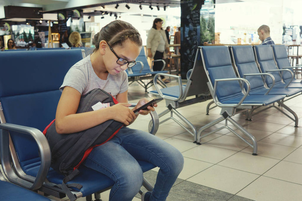 Χαριτωμένο κορίτσι με τα γυαλιά είναι κάθεται στο αεροδρόμιο σε αναμονή για την πτήση και να ψάχνει σε smartphone. - Φωτογραφία, εικόνα