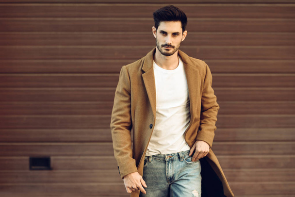 Young man wearing winter clothes in the street. Young bearded guy with modern hairstyle with coat, blue jeans and white t-shirt. - Photo, Image