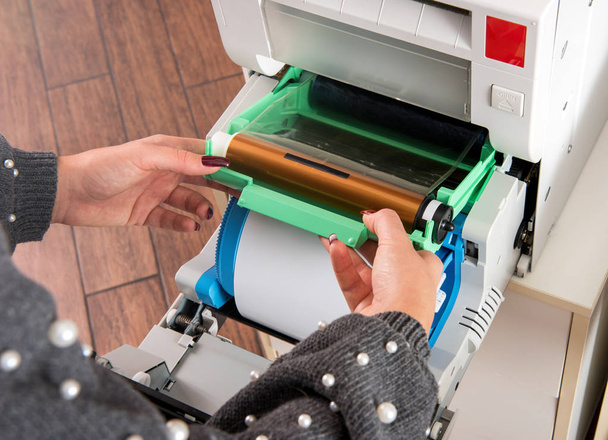 Woman changing the ribbon on a photographic printer above a new roll of film paper in a close up view of the machine and her hands - Photo, Image