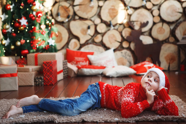 Chłopiec w Swiąteczne są kłamstwa i śmiejąc się na dywanie w pobliżu drzewa nowy rok w tle kawałki drzewa. - Zdjęcie, obraz
