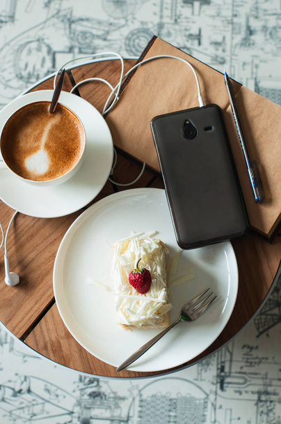 Cup of coffee, smart phone and headphones, open notebook with pen, delicious cake with chocolate and strawberry in a plate. Top view. Life style. Flat lay - Photo, Image