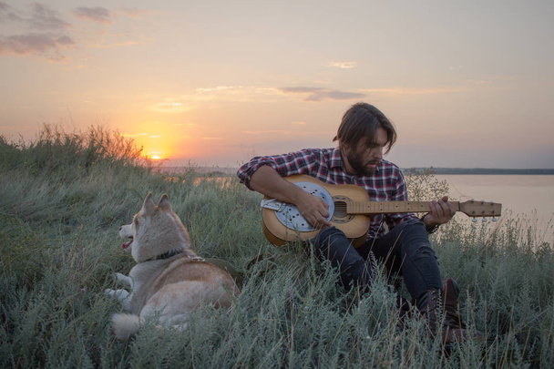 young handsome men on the cliff with guitar and husky dog during sunset in lake - Zdjęcie, obraz