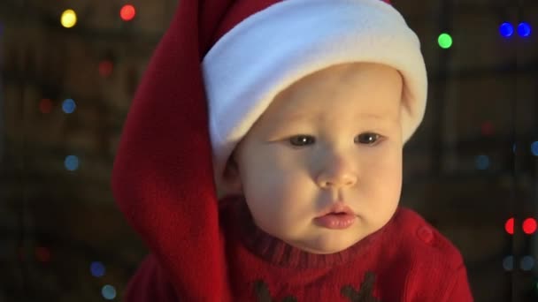 Cute little child in Santa hat over Christmas lights background. New Year celebration concept. - Кадры, видео
