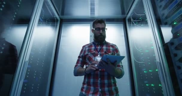 Wide shot van casual man met tablet wandelen onder server racks in data center kamer kijken naar hardware - Video