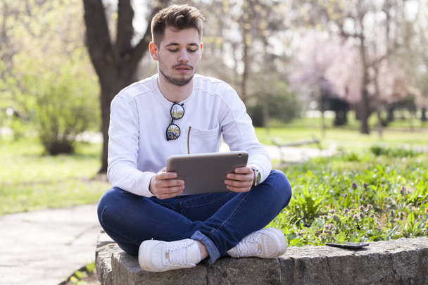 Yakışıklı genç adam parkta oturan tablet kullanmak ve açık havada. Seçici odak ve küçük alan derinliği. - Fotoğraf, Görsel