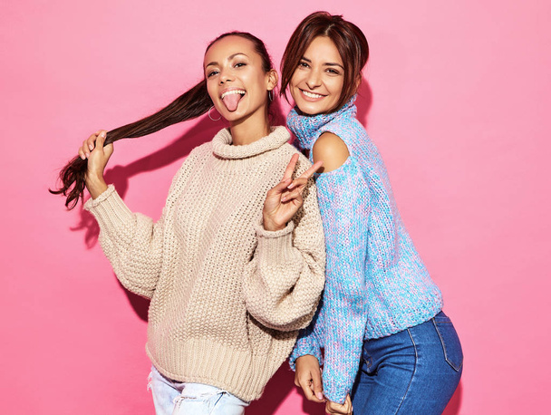 Two beautiful sexy smiling gorgeous girls looking at camera. Hot women standing in stylish white and blue sweaters, on pink background. Showing peace sign - Photo, image