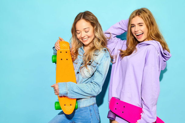 Two young stylish smiling blond women with penny skateboards. Girl in summer hipster sport clothes posing near blue wall in studio. Positive models - Фото, изображение
