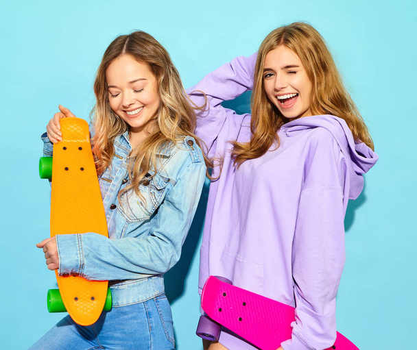 Two young stylish smiling blond women with penny skateboards. Girl in summer hipster sport clothes posing near blue wall in studio. Positive models - Foto, Imagem