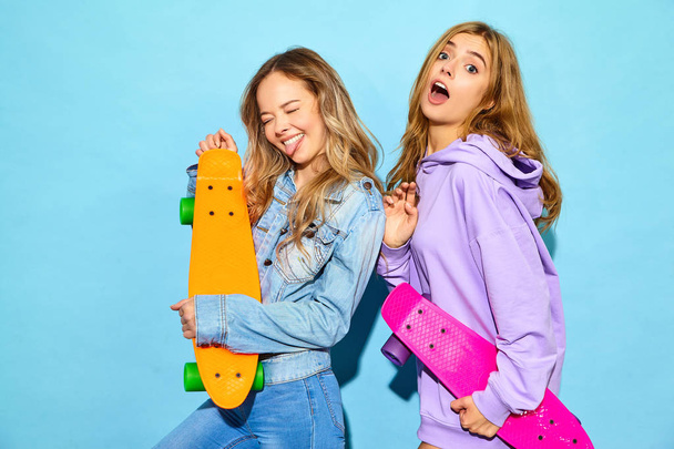 Two young stylish smiling blond women with penny skateboards. Girl in summer hipster sport clothes posing near blue wall in studio. Positive models - 写真・画像