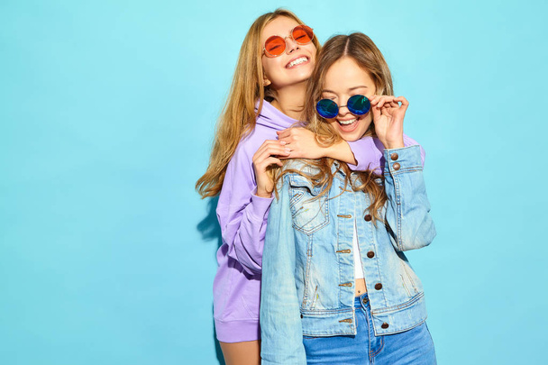 Two young beautiful blond smiling hipster girls in trendy summer clothes. Sexy carefree women posing near blue wall in sunglasses. Positive models going crazy and hugging - Fotoğraf, Görsel